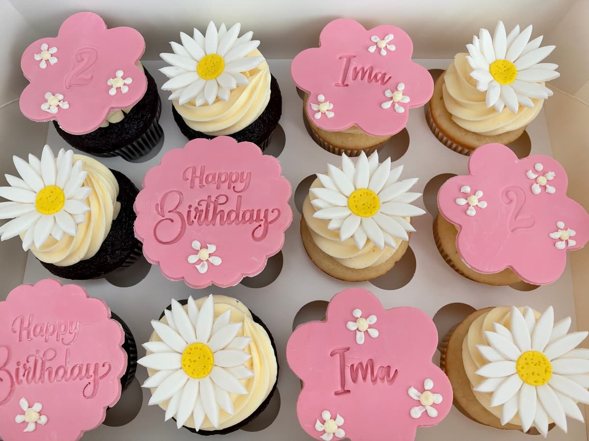 Box of chocolate and vanilla birthday cupcakes with fondant daisies and pink flower-shaped Happy Birthday, name, and number toppers